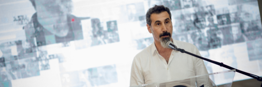 Serj Tankian standing at a podium with his hands in his pockets in front of a presentation slide