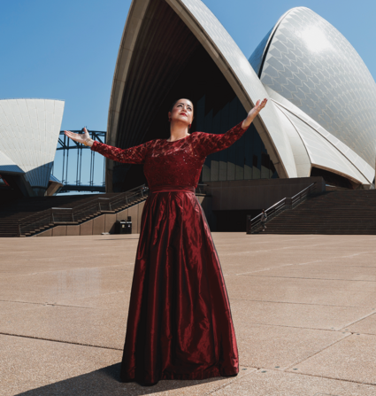 Soprano woman in a red dress stands with her arms open at the Opera House in Sidney, Australia