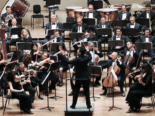 A variety of musicians dressed in black (the Armenian National Philharmonic Orchestra) playing instruments and being conducted front and center by Eduard Topchjan. They are performing in Beijing, China in 2019.