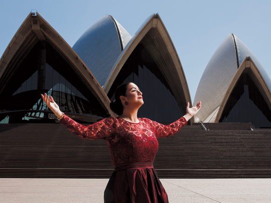 Soprano woman in a red dress stands with her arms open at the Opera House in Sidney, Australia