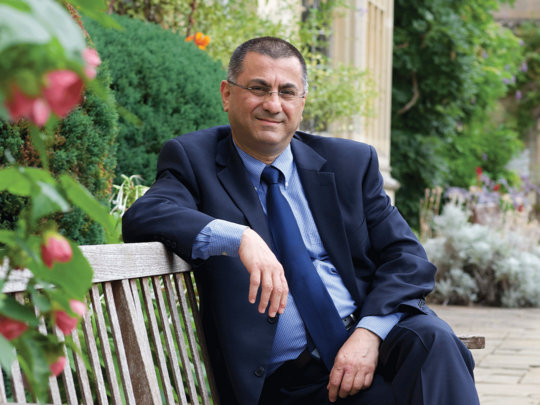 University professor with glasses and blue suit poses in a garden at University of Oxford campus